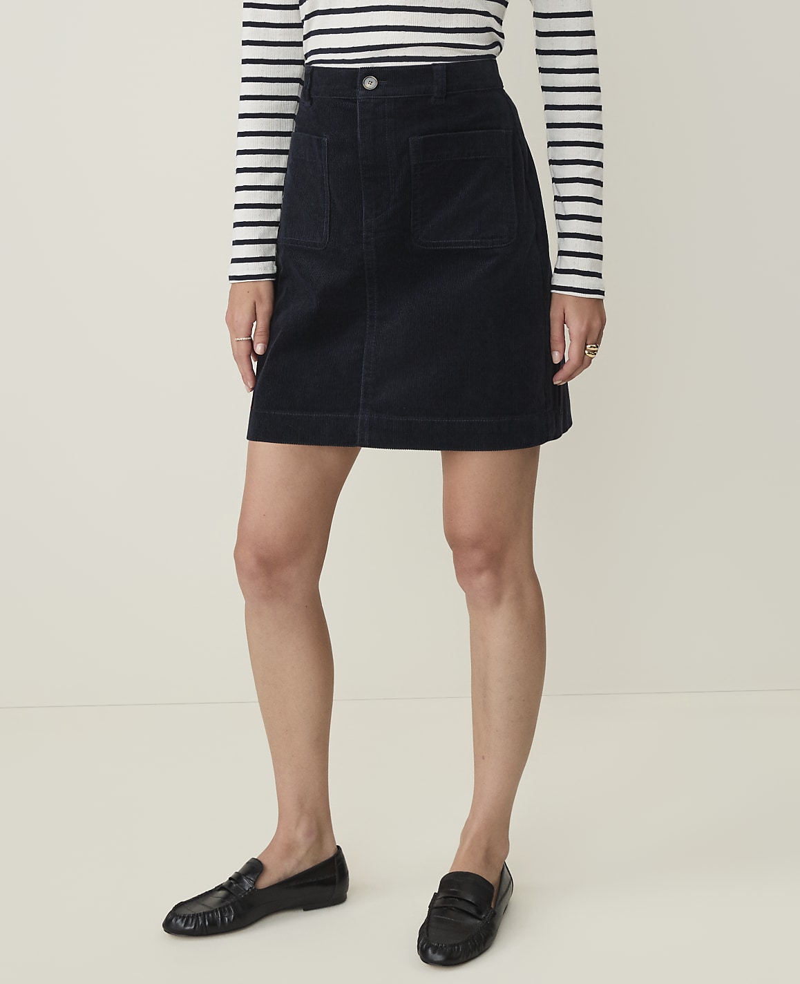 A woman in a navy corduroy skirt and striped shirt stands against a plain, light-colored background.