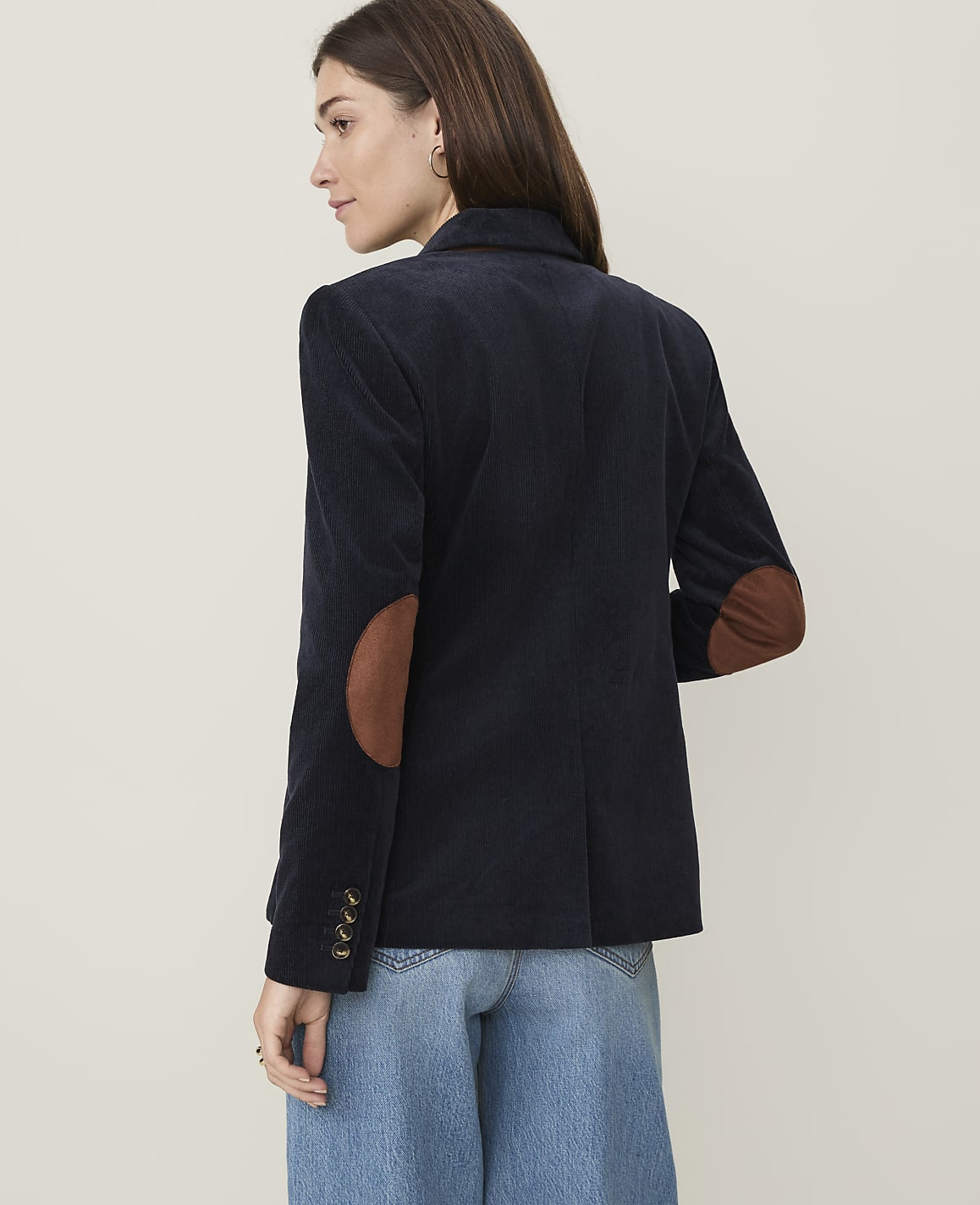 A woman in a navy corduroy blazer with suede elbow patches stands against a plain, light-colored background.