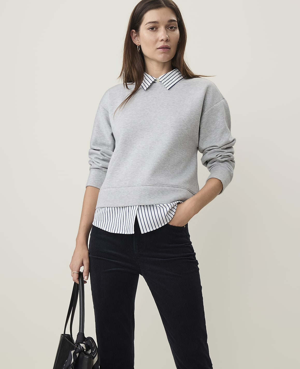A woman in a grey sweatshirt layered over a striped collared shirt and dark pants stands against a plain, light background.