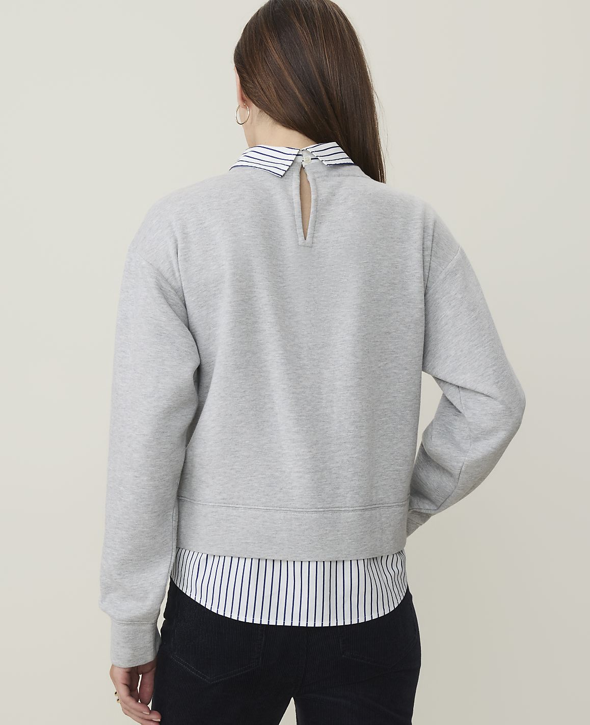 A woman wearing a grey sweatshirt layered over a striped collared shirt stands against a plain, light-colored background.