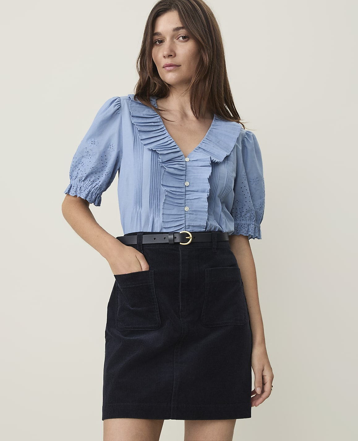A woman in a blue ruffled blouse and dark skirt stands against a plain, light background.