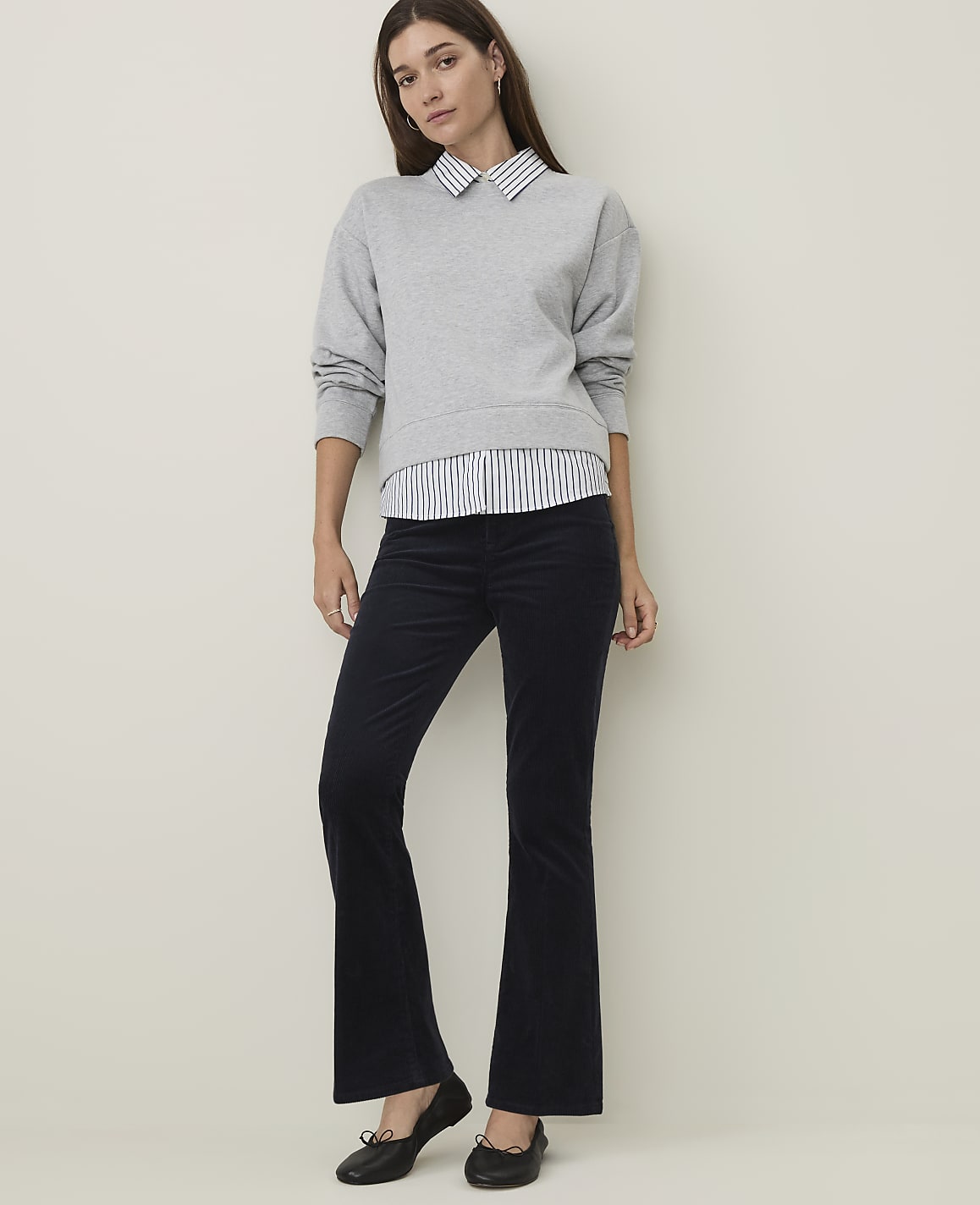 A woman in a grey sweatshirt layered over a striped shirt and dark flared pants stands against a plain, light-colored wall.