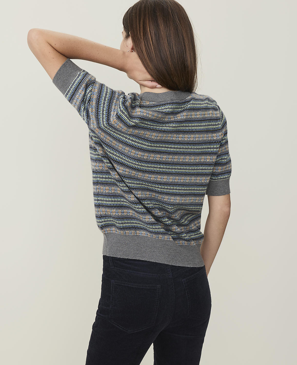 A woman in a striped sweater and dark pants stands against a plain, light-colored background.