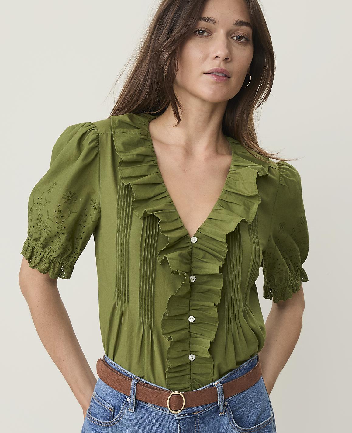 A woman in a ruffled green blouse and blue jeans stands against a plain, light background.