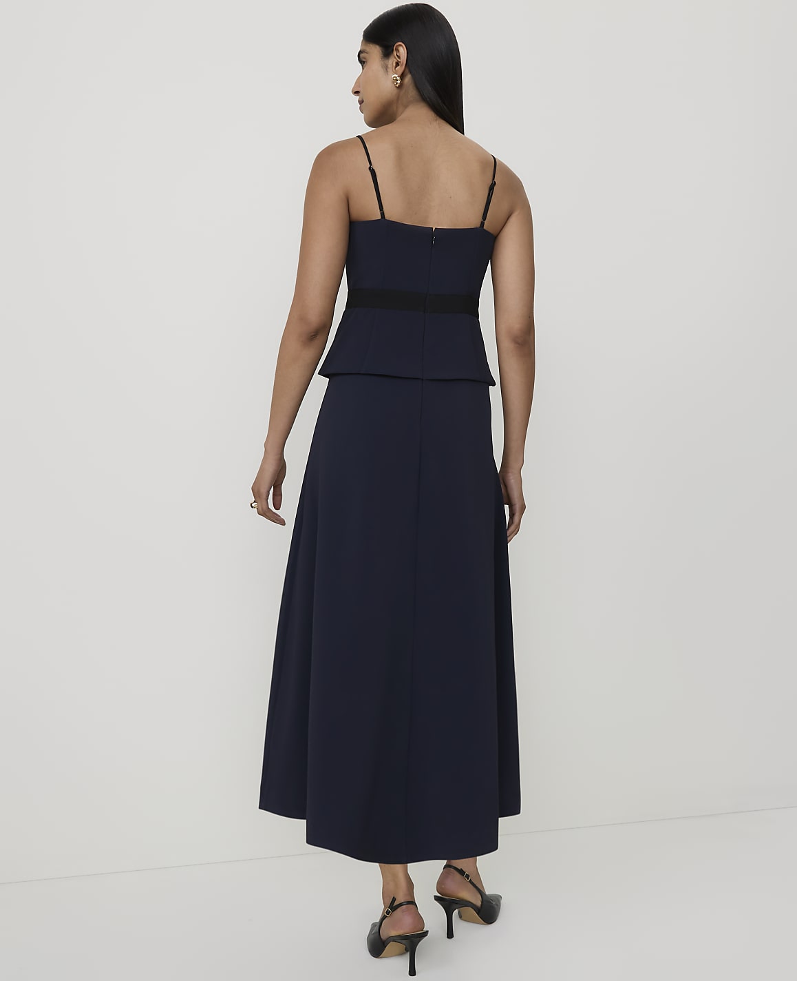 A woman in a navy blue dress with spaghetti straps and a black waistband stands against a plain white background.