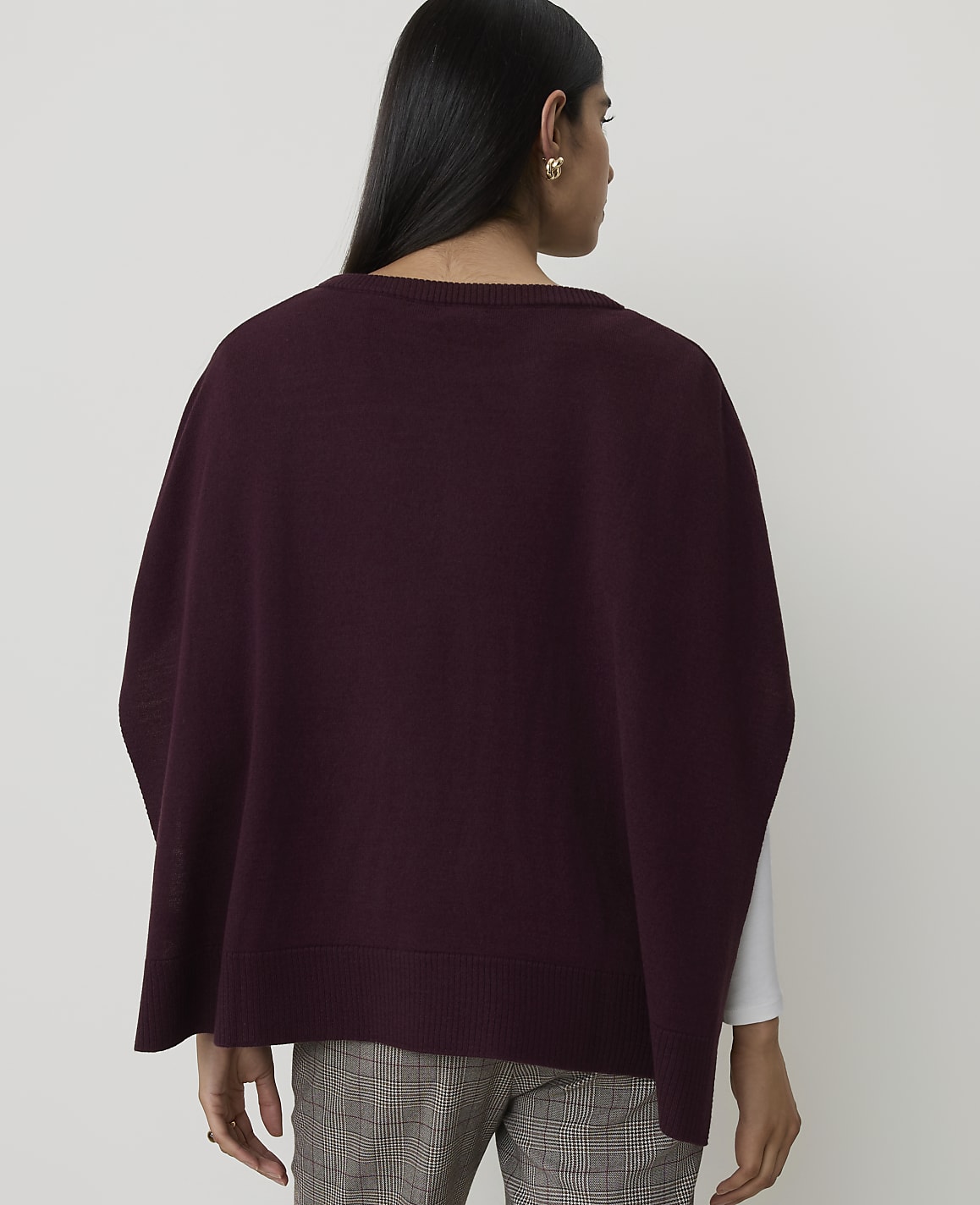A woman wearing a burgundy poncho over a white shirt and plaid pants stands against a plain white background.