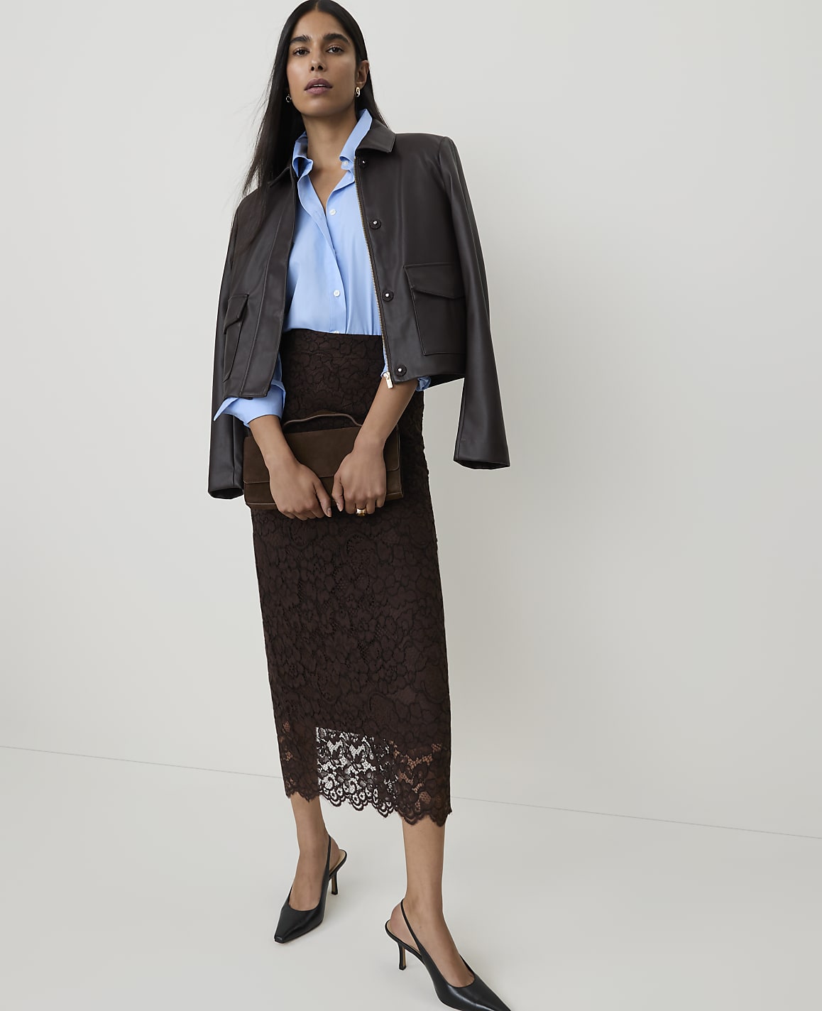 A woman in a brown lace skirt and a blue shirt with a leather jacket draped over her shoulders stands against a plain white background.