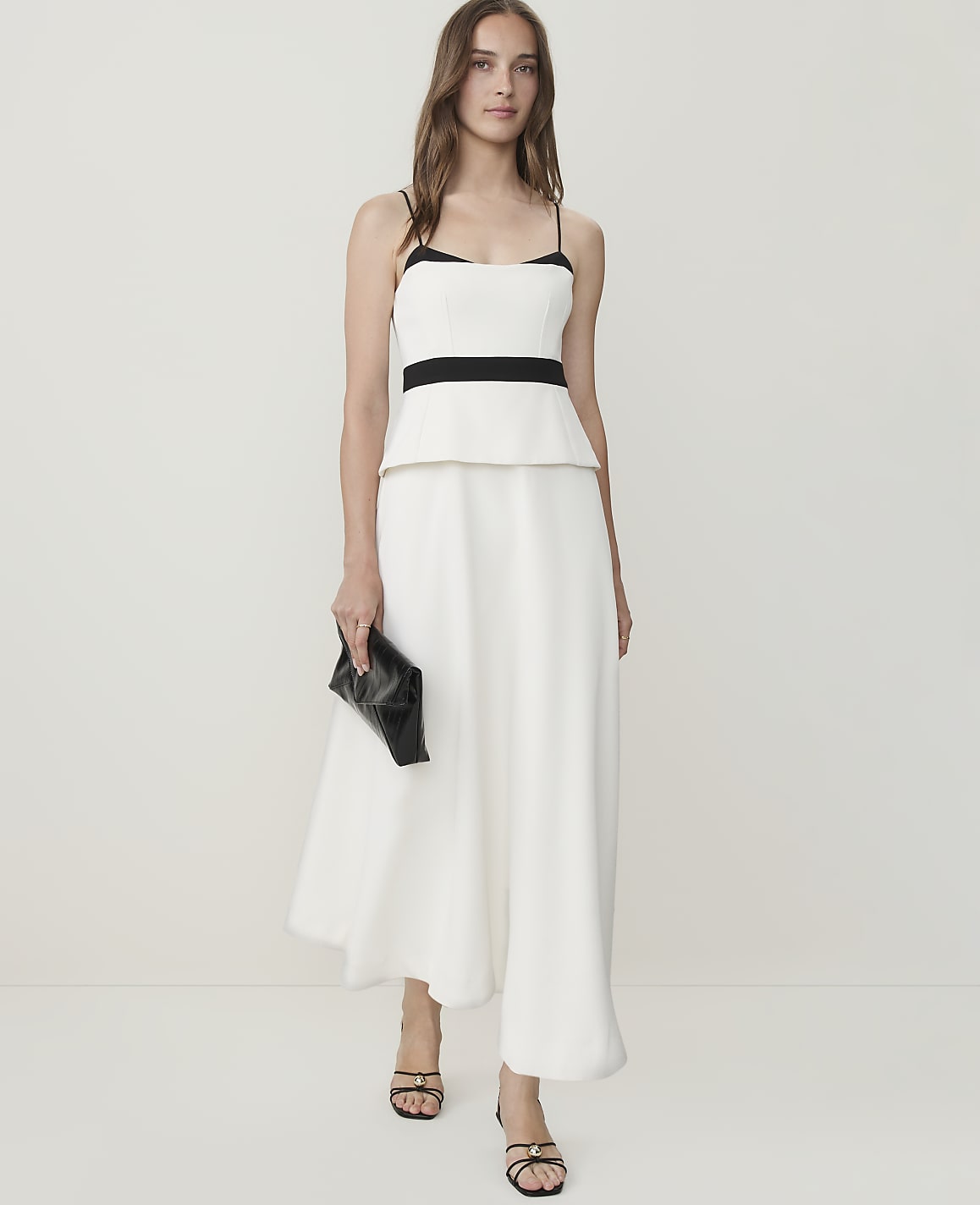 A woman in a white peplum top and wide-leg pants holds a black clutch against a plain white background.
