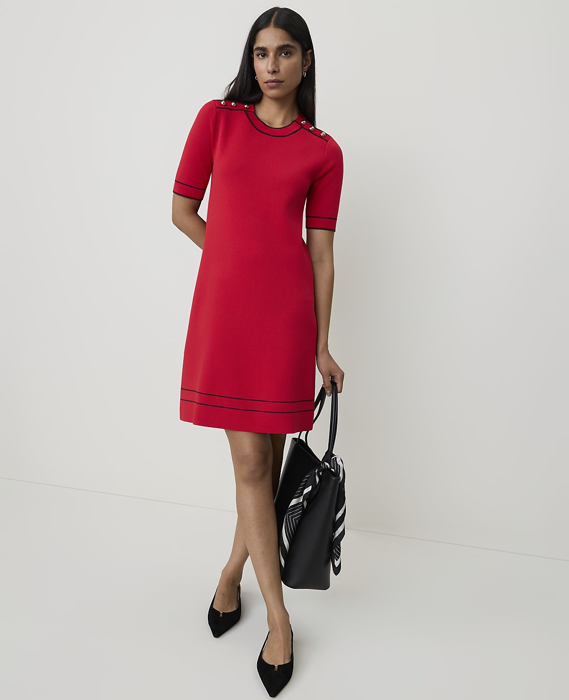 A woman in a red dress stands against a plain white background, holding a black tote bag.