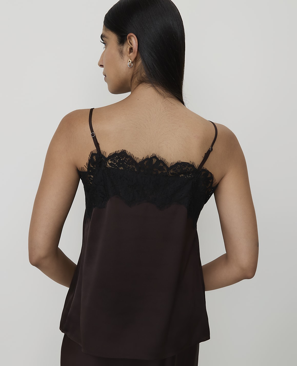 A woman in a brown camisole with black lace trim stands against a plain white background.