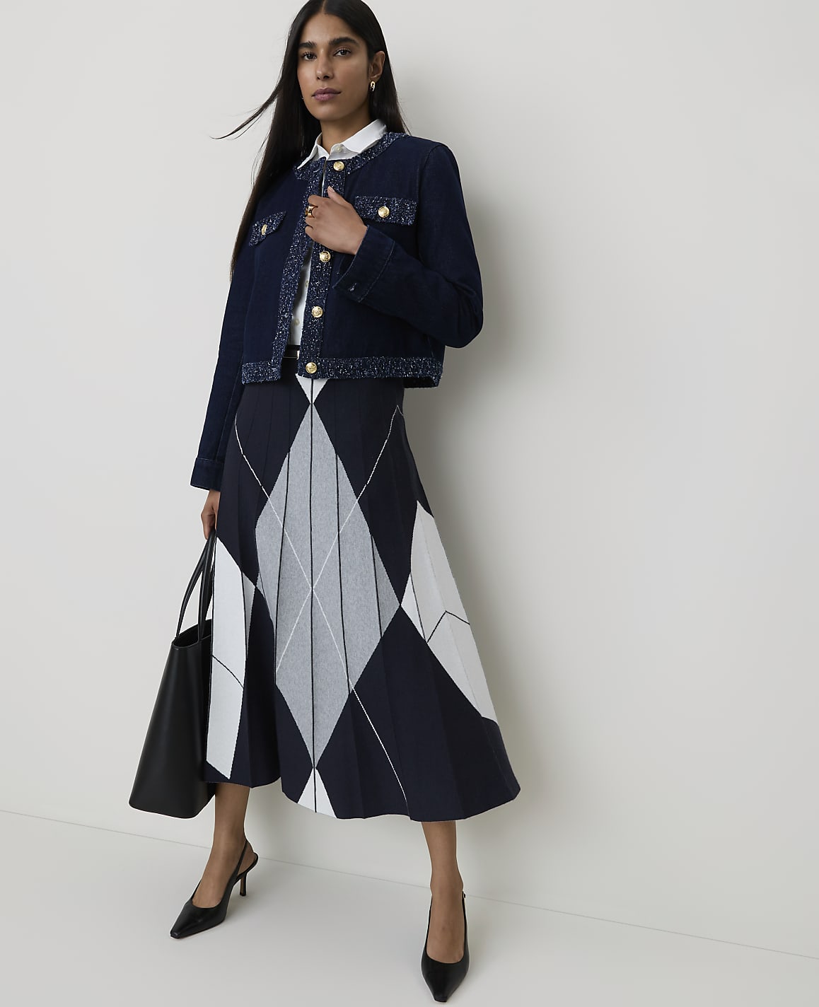 A woman in a dark blue tweed jacket and a patterned skirt stands against a plain white background, holding a black tote bag.