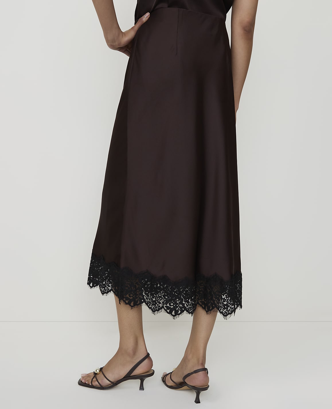 A person stands in front of a plain white background, wearing a brown satin skirt with a black lace hem and strappy sandals.