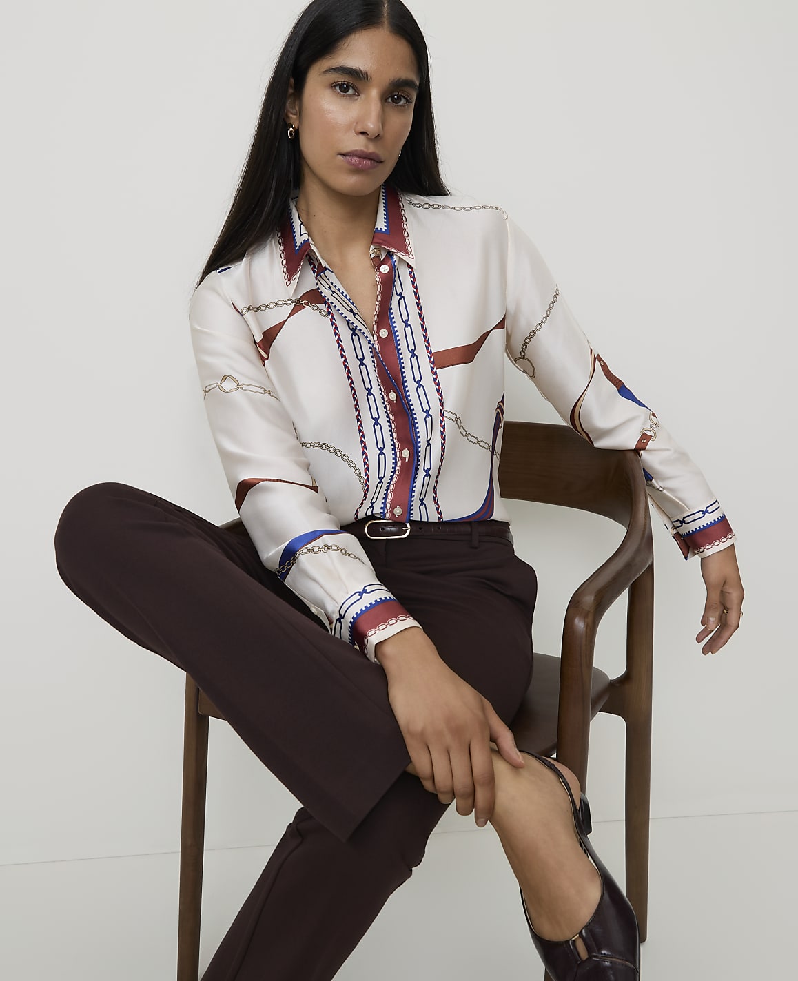 A woman in a patterned silk shirt and dark trousers sits on a wooden chair against a plain background.