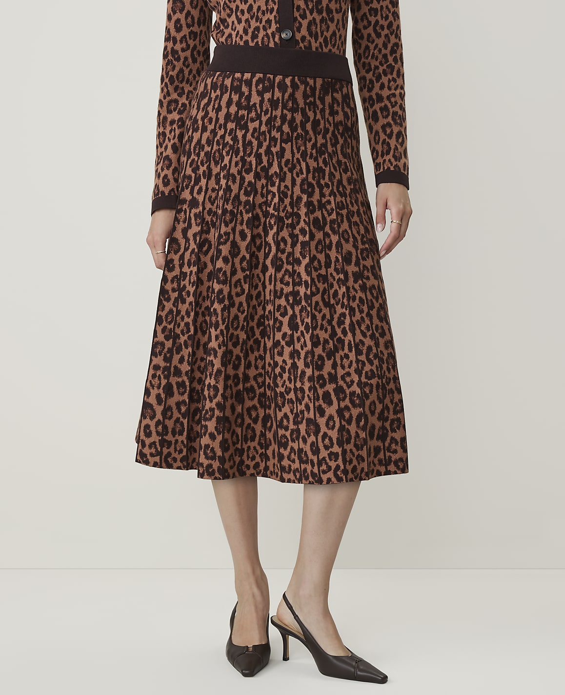 A person stands in a studio wearing a leopard print pleated skirt and matching cardigan, with a plain background.