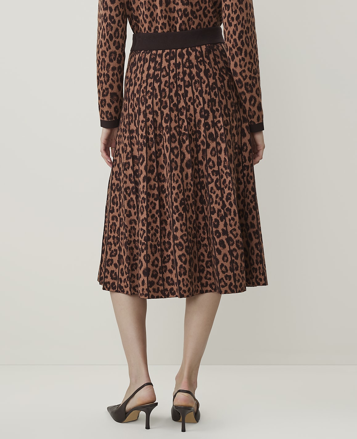 A person wearing a brown leopard-print pleated skirt and a matching top stands against a plain, light-colored background.