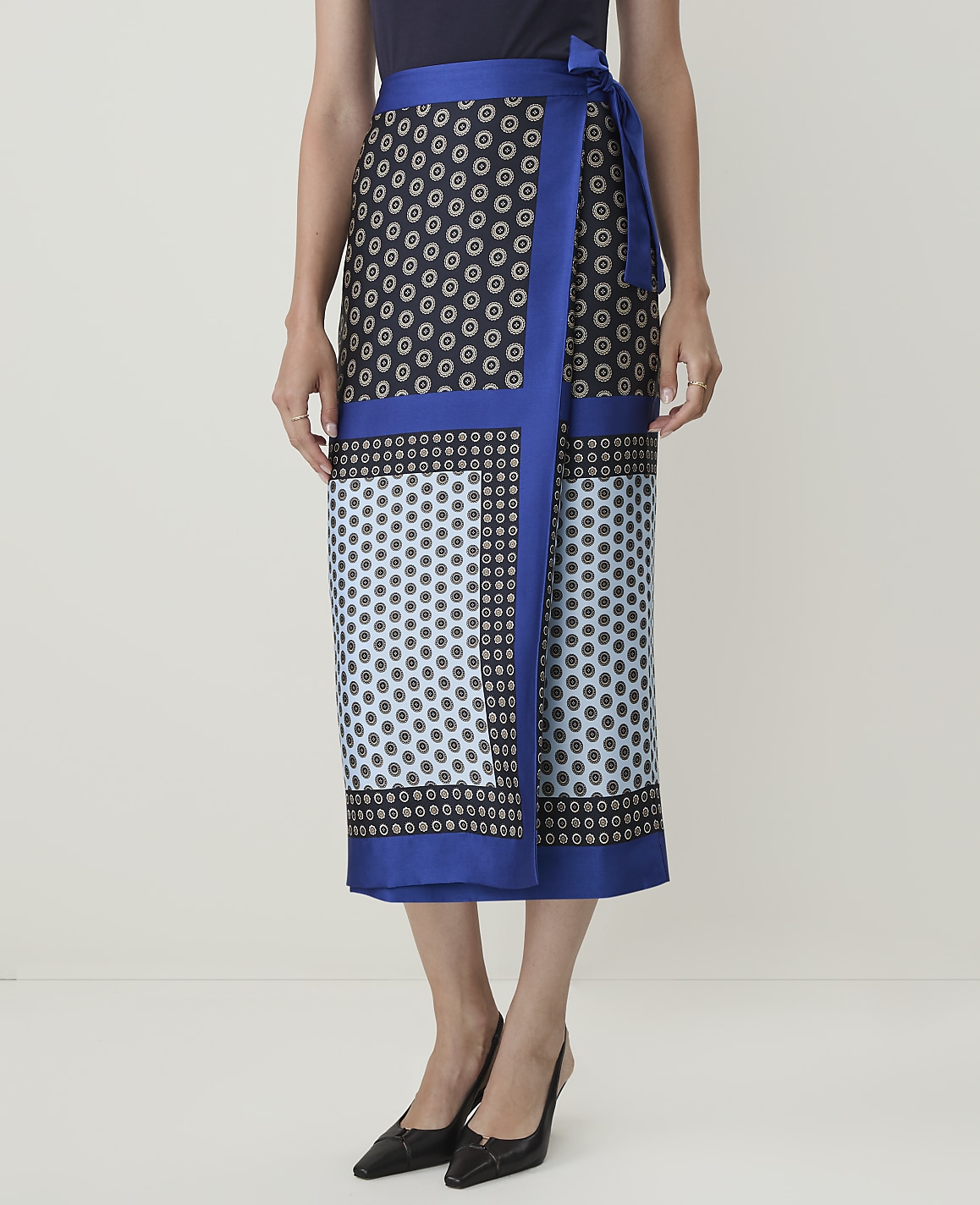 A woman models a patterned wrap skirt with a blue border, standing against a plain, light-colored background.
