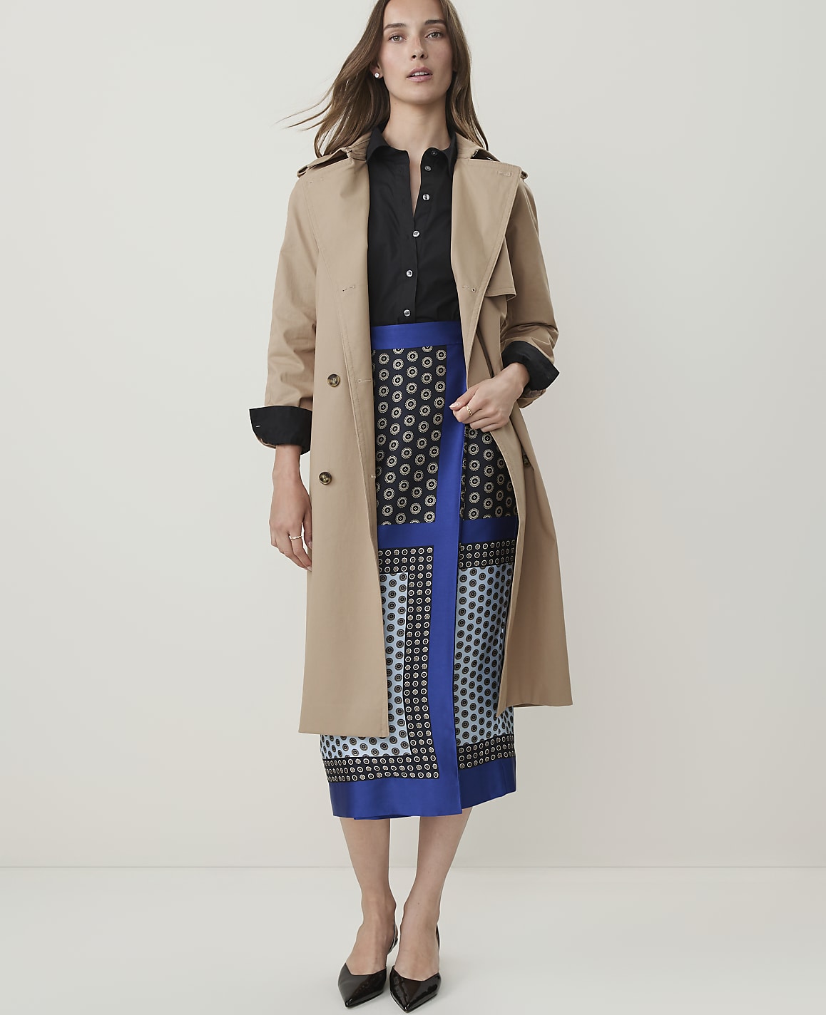 A woman in a trench coat and patterned skirt stands against a plain, light-colored background.
