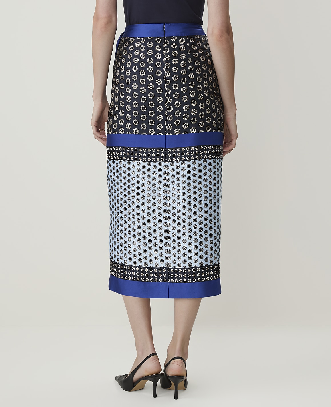 A woman in a patterned skirt stands against a plain, light-colored background.