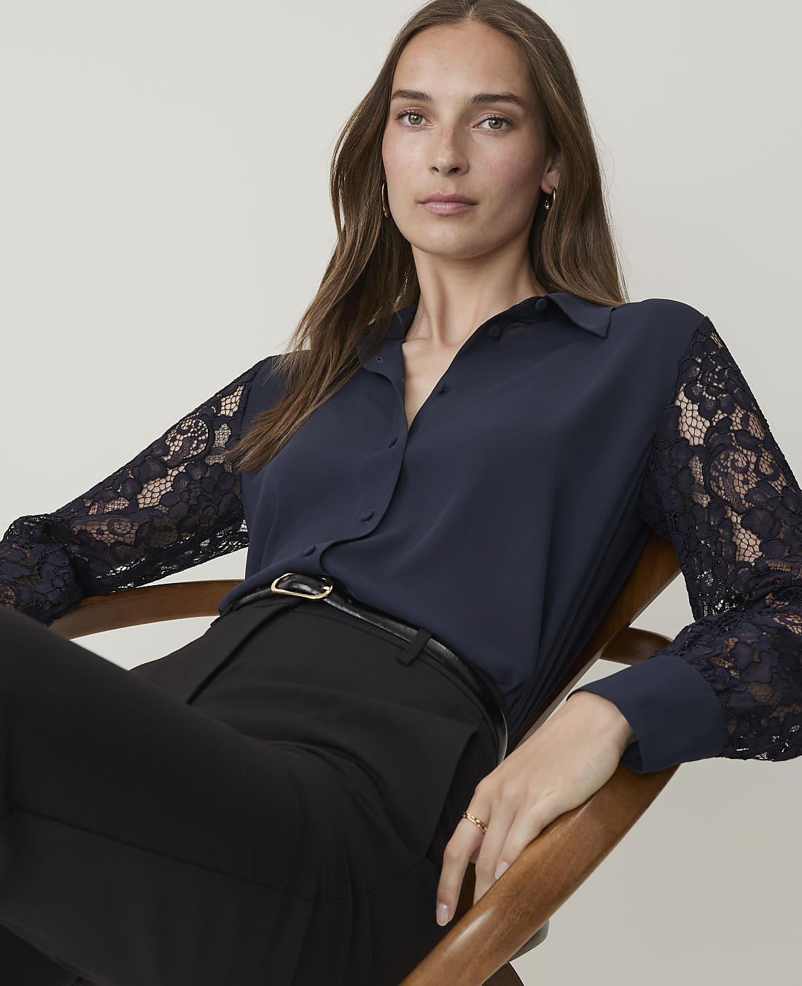 A woman in a navy blue blouse with lace sleeves and black pants sits in a chair against a plain background.
