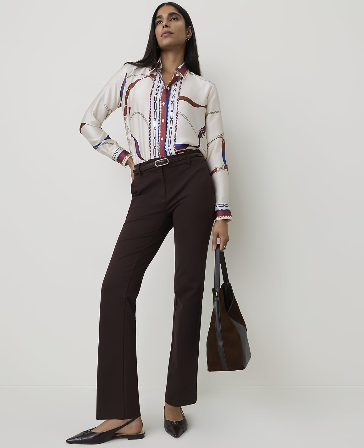 A woman in a patterned blouse and brown pants stands against a plain white background, holding a dark brown bag.