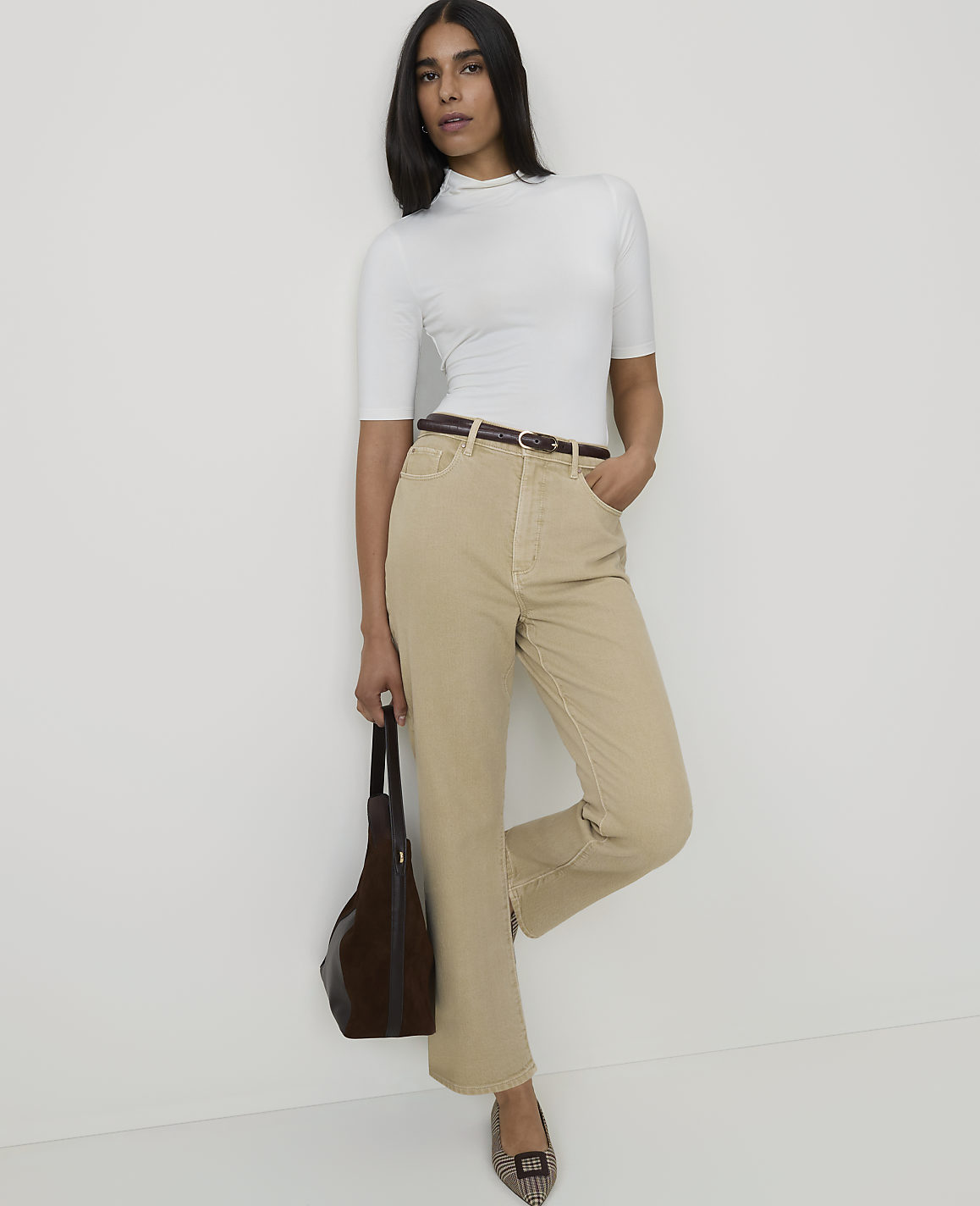 A woman in a white turtleneck and tan pants stands against a plain white background, holding a brown purse.