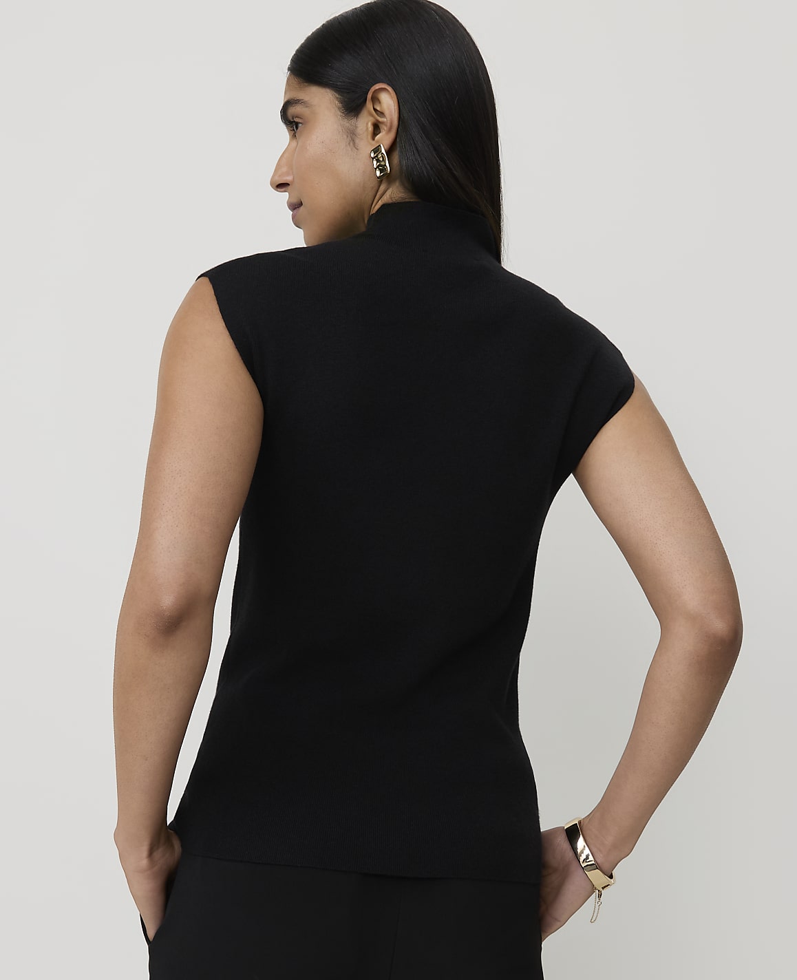 A woman in a black sleeveless turtleneck stands with her back to the camera, against a plain white background.