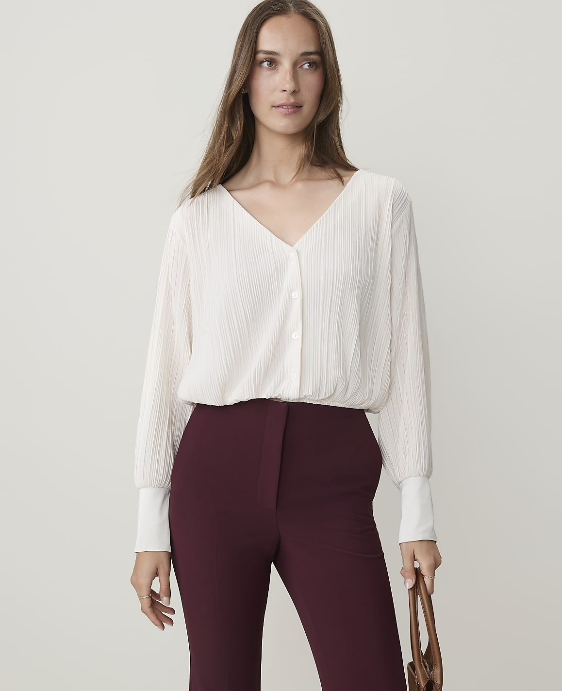 A woman in a cream-colored blouse and burgundy pants stands against a plain, light background, holding a brown bag.