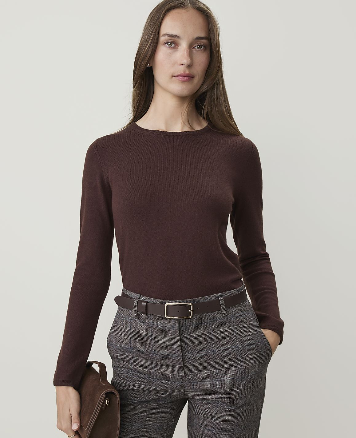A woman in a brown sweater and plaid pants stands against a plain background, holding a brown clutch.