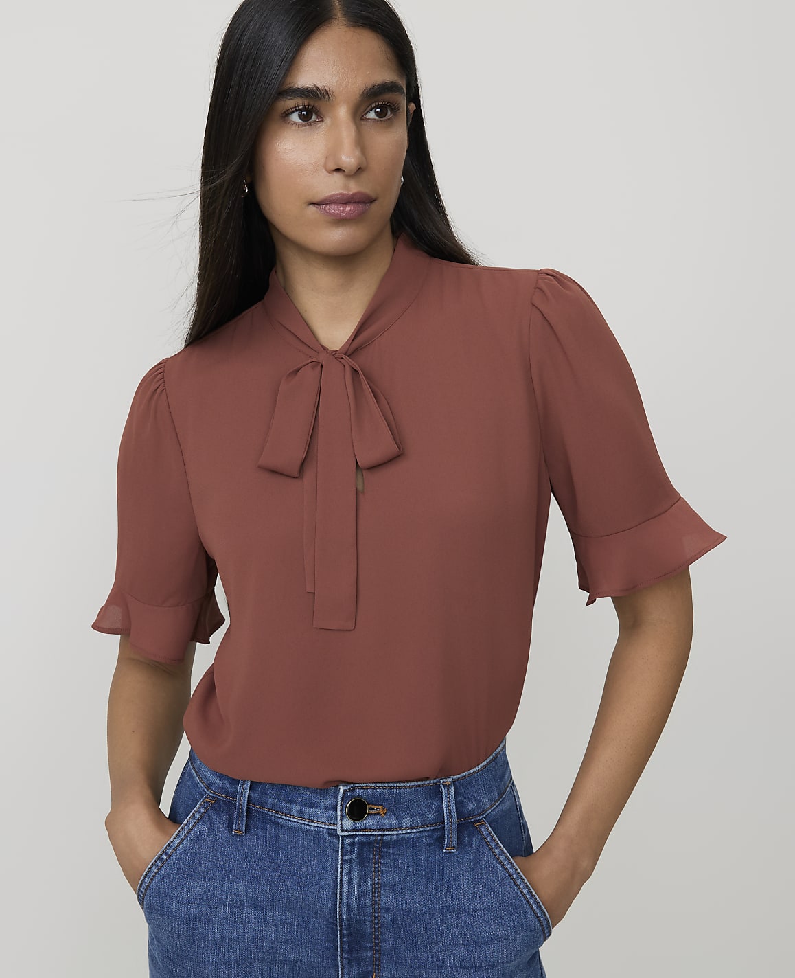 A woman in a rust-colored bow-neck blouse and blue jeans stands against a plain white background.