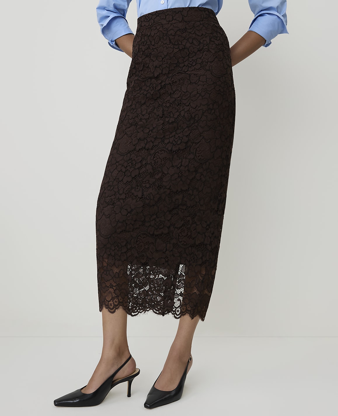 A woman in a brown lace skirt and blue shirt stands against a plain white background, wearing black slingback heels.