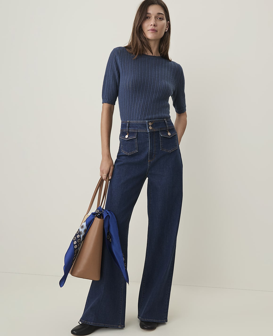 A woman in a blue ribbed sweater and dark wash wide-leg jeans stands holding a brown tote bag with a blue scarf, against a plain, light background.