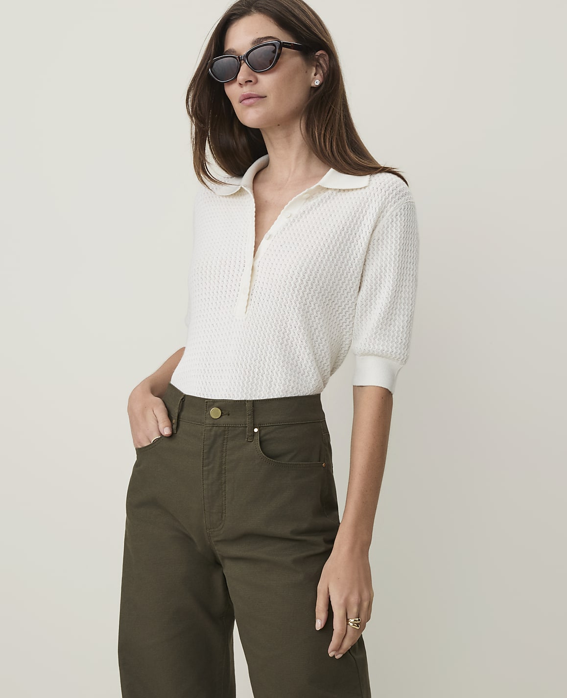 A woman in a white polo shirt and olive green pants stands against a plain, light background.