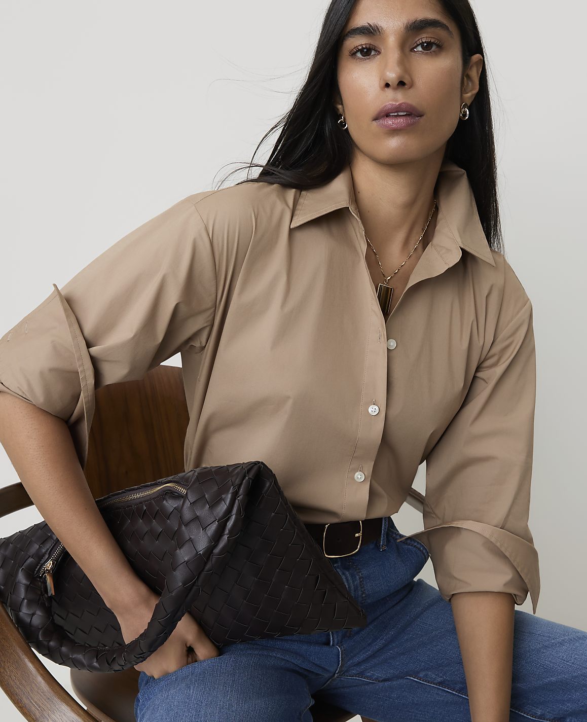 A woman in a tan button-down shirt and jeans holds a dark woven clutch in front of a plain background.