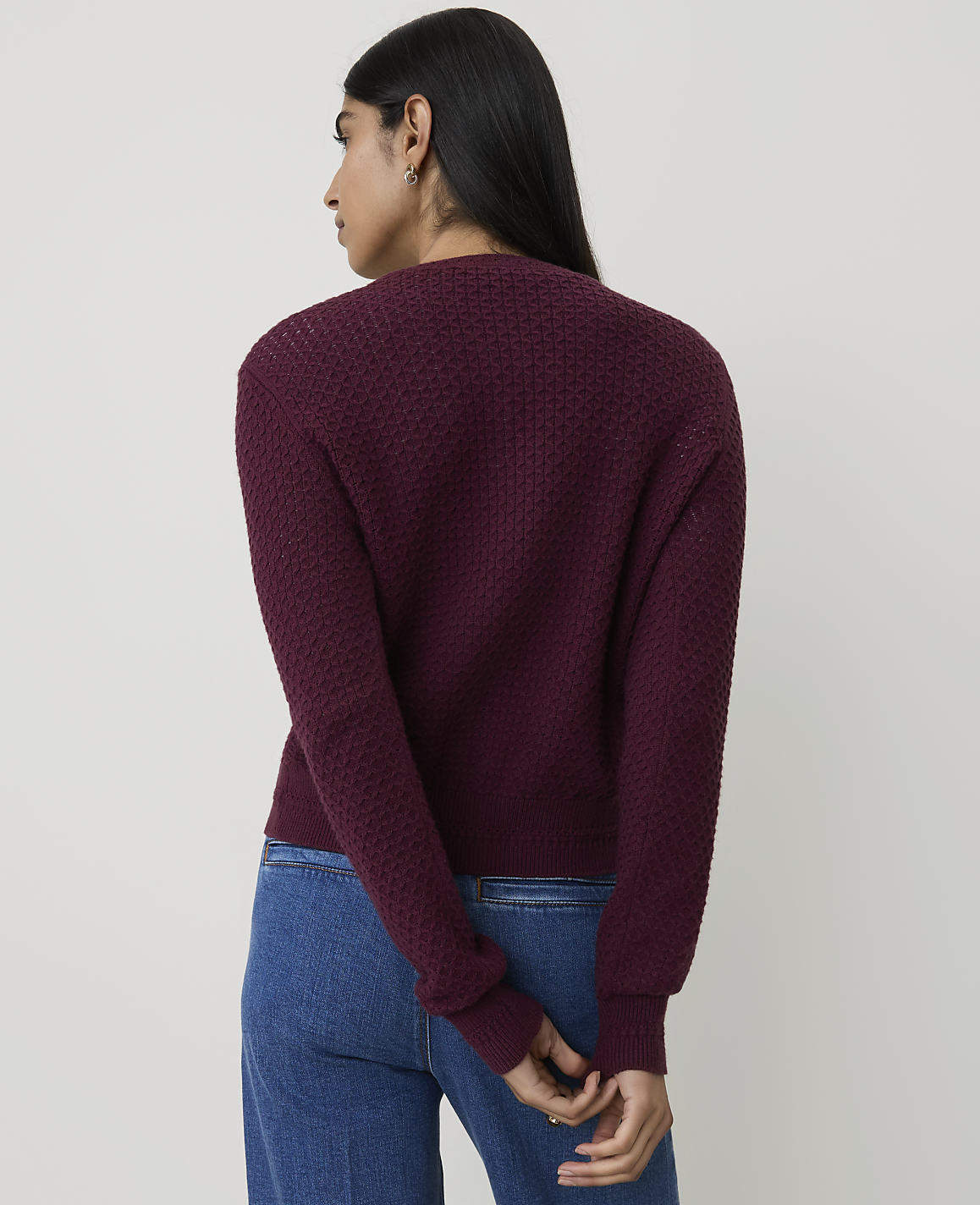A woman in a maroon textured sweater and blue jeans stands against a plain, light-colored background.