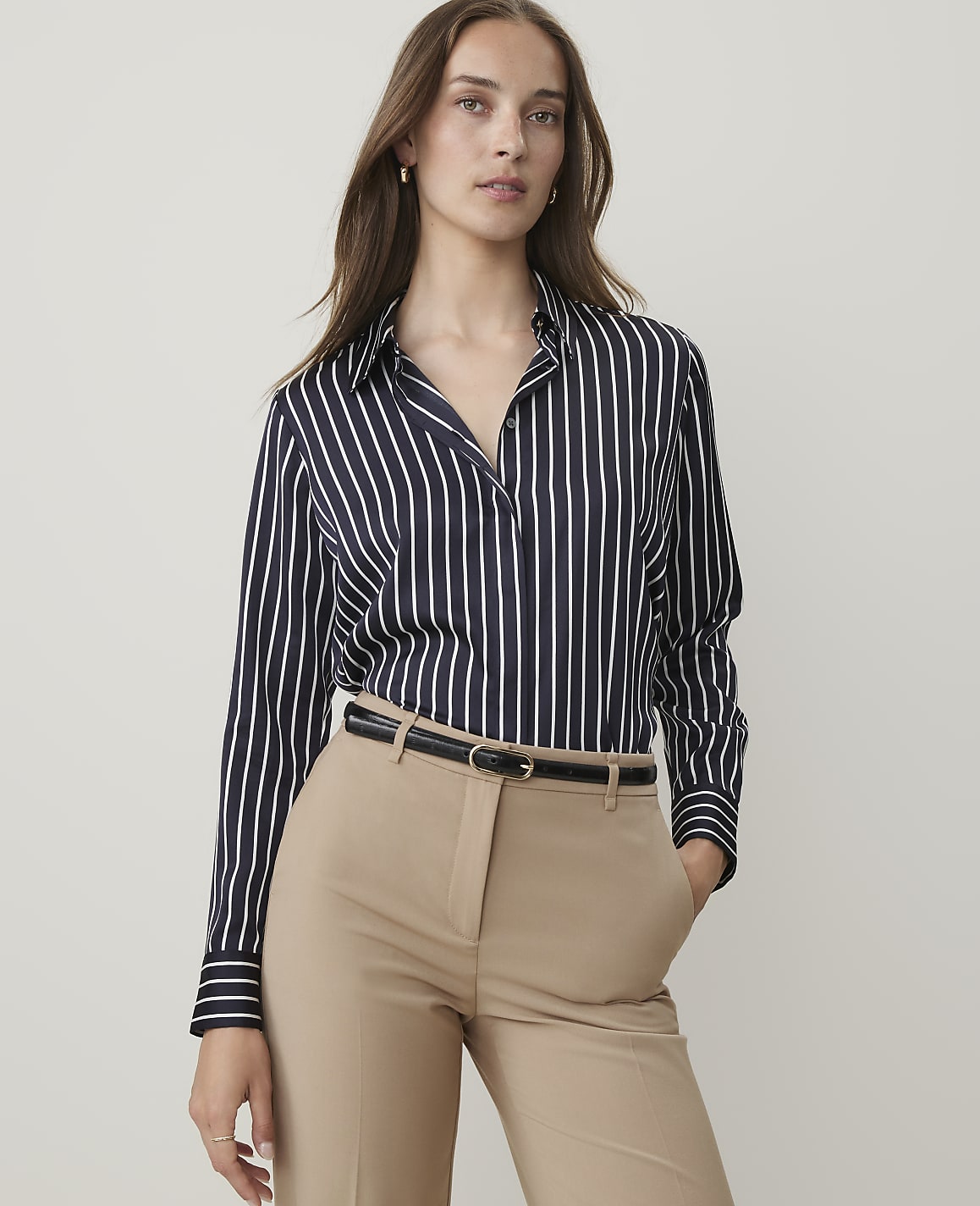 A woman in a striped blouse and tan pants stands against a plain background.