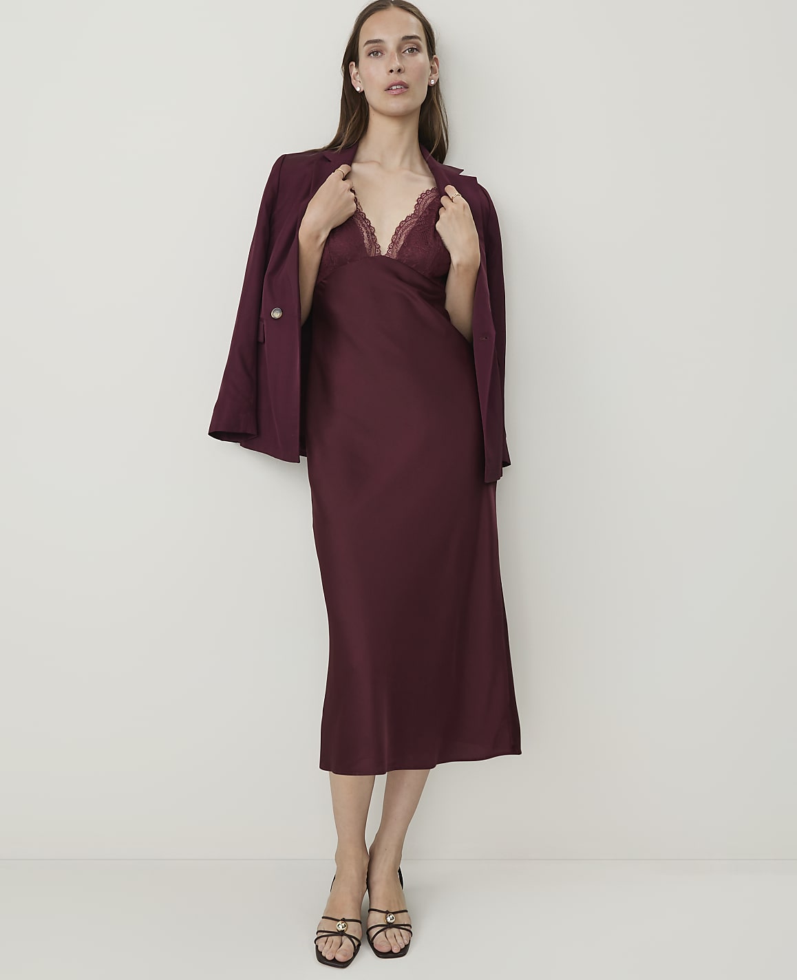 A woman in a burgundy slip dress and blazer stands against a plain white background.