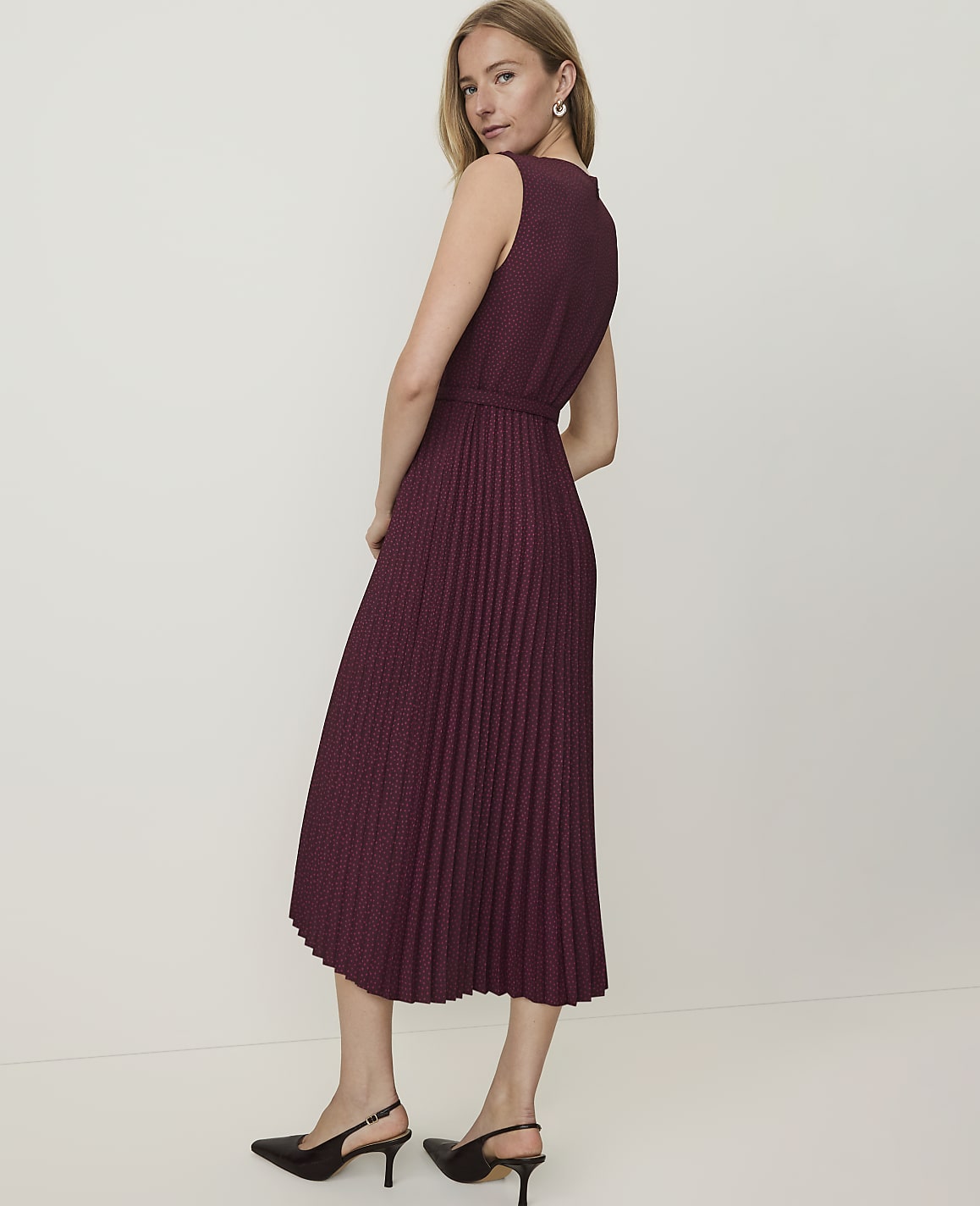 A woman in a burgundy pleated dress and black heels stands against a plain white background.