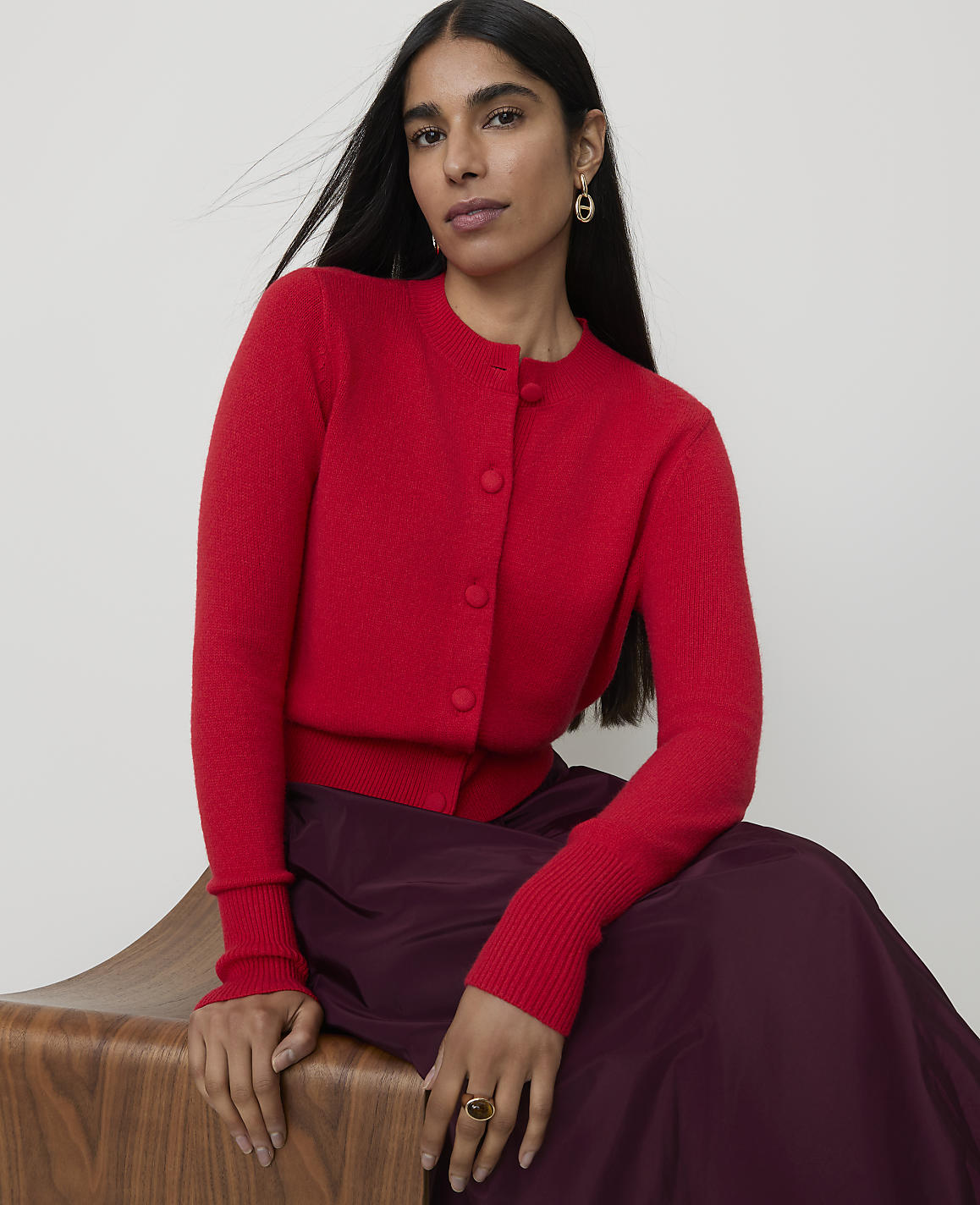 A woman in a red cardigan and plum skirt sits on a wooden stool against a plain background.