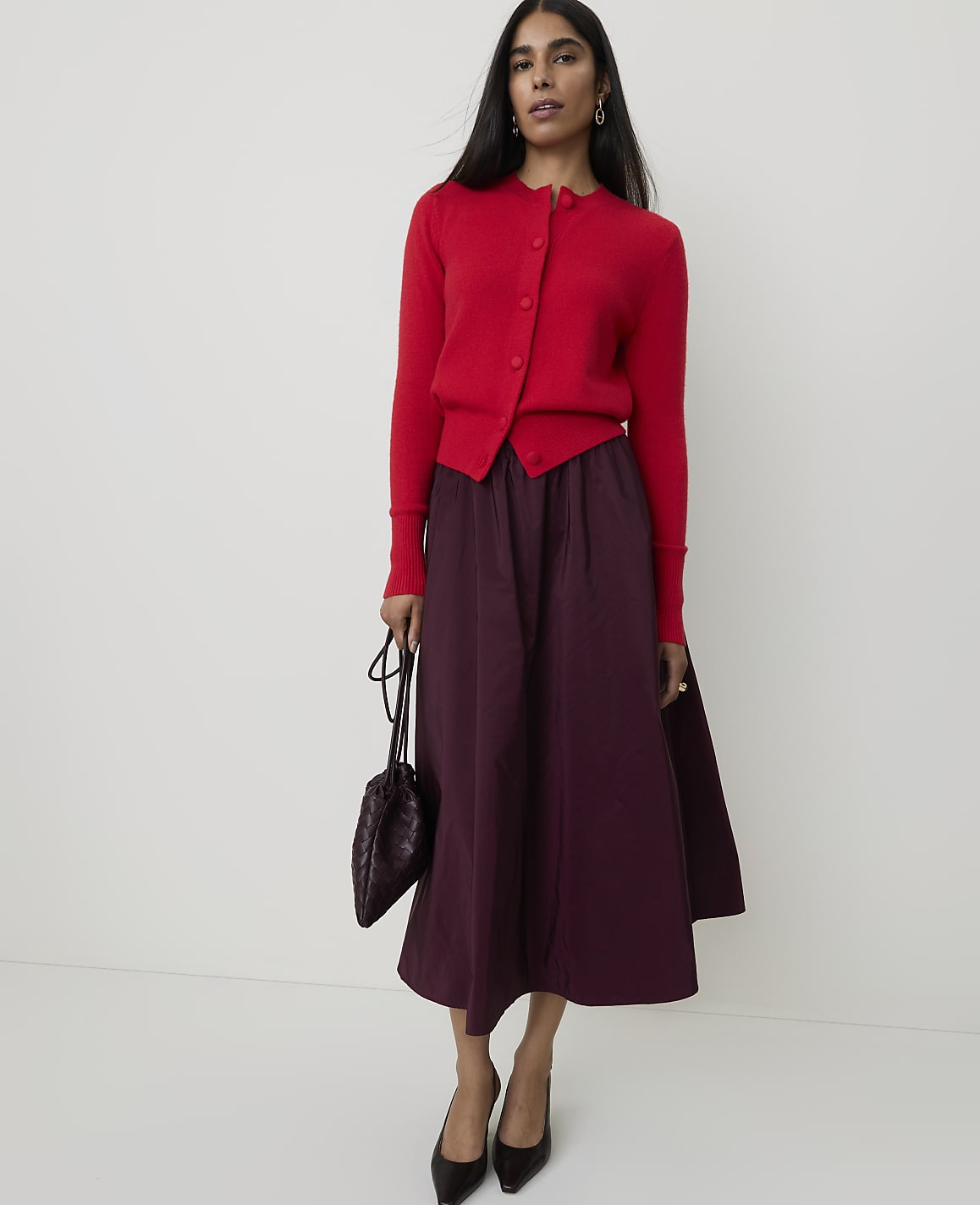 A woman in a red cardigan and plum skirt stands against a plain white background, holding a dark patterned handbag.