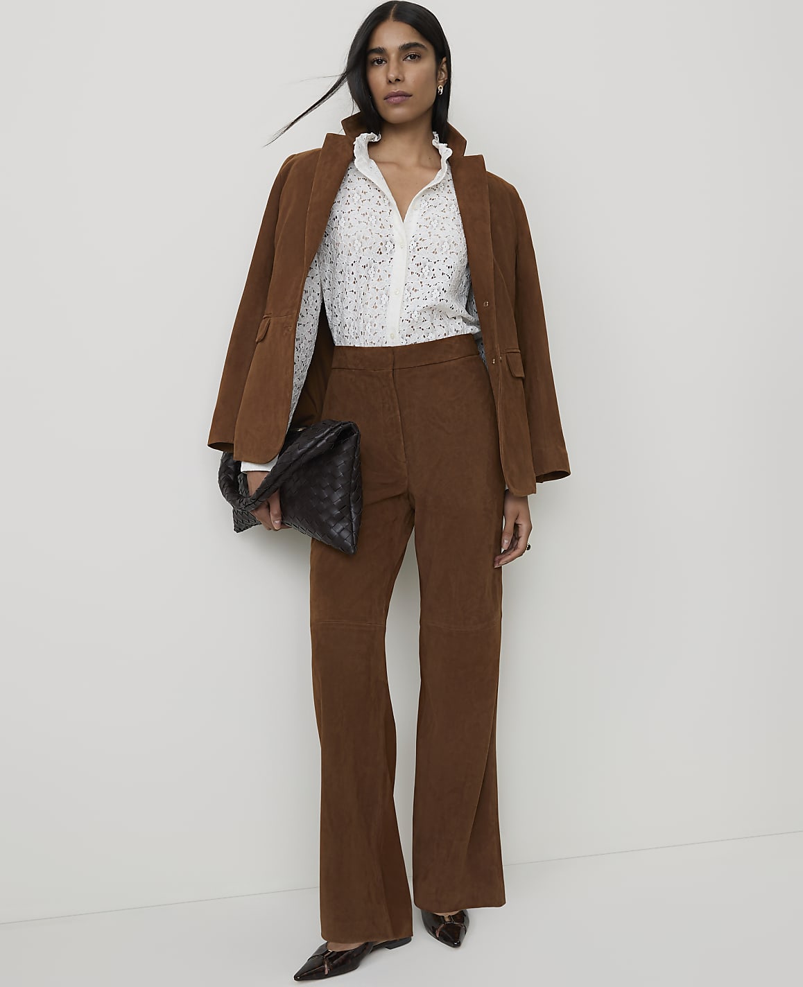 A woman in a brown corduroy suit and a patterned blouse stands against a plain white background, holding a black woven clutch.