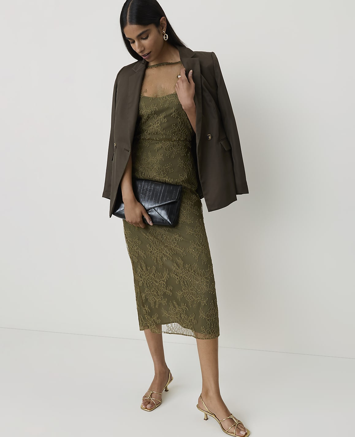 A woman in a green lace dress and brown blazer holds a black clutch while standing against a plain white background.