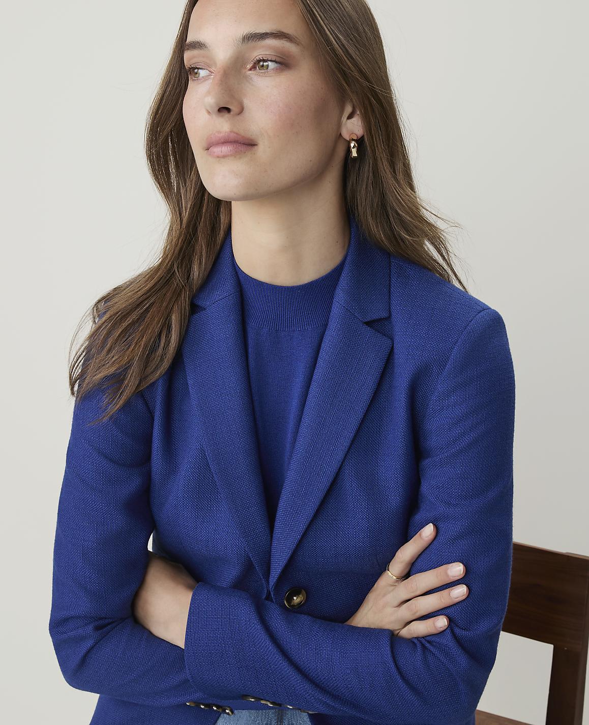 A woman in a blue blazer with her arms crossed is looking away from the camera against a plain, light-colored background.