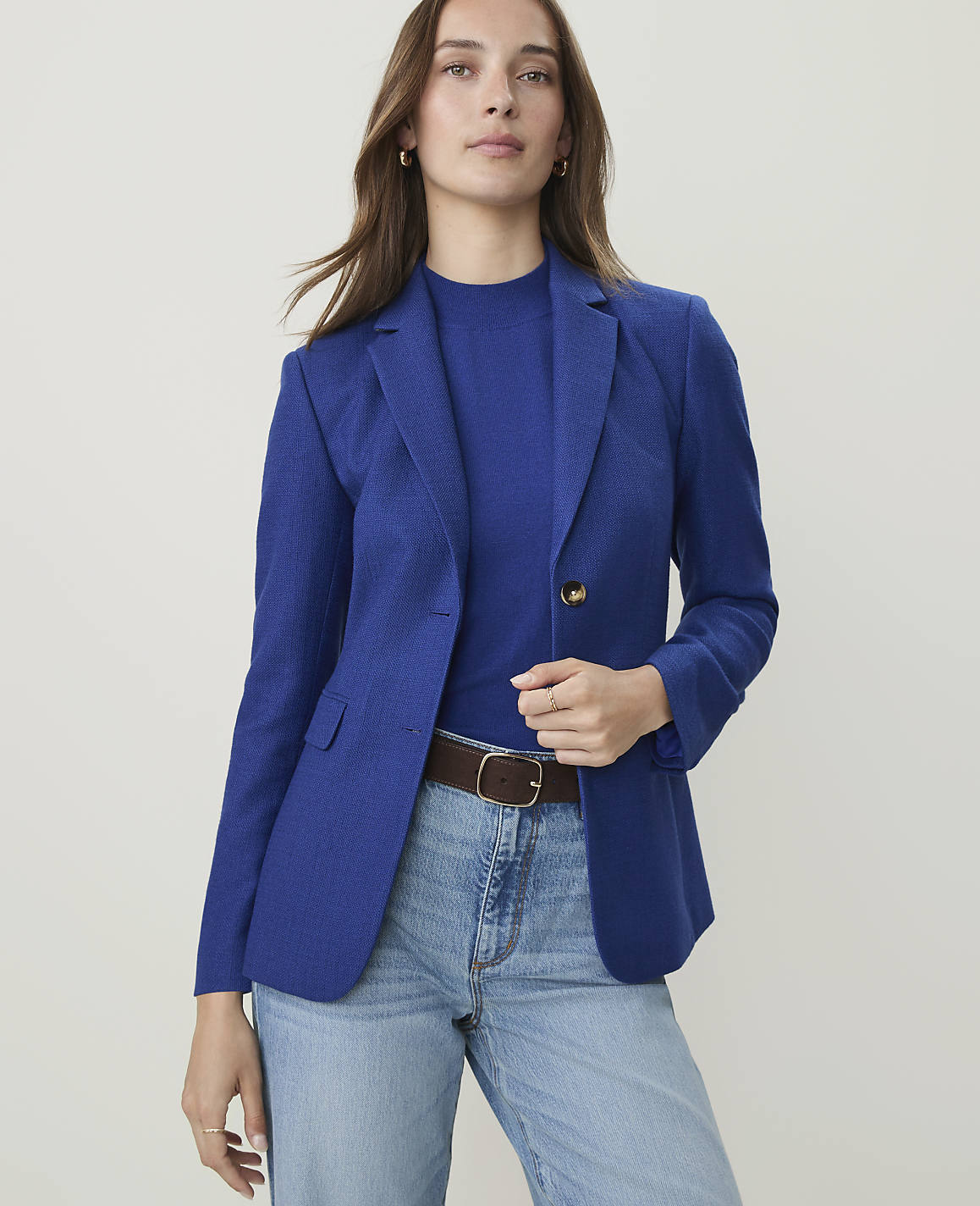 A woman in a blue blazer and jeans stands against a plain, light background.