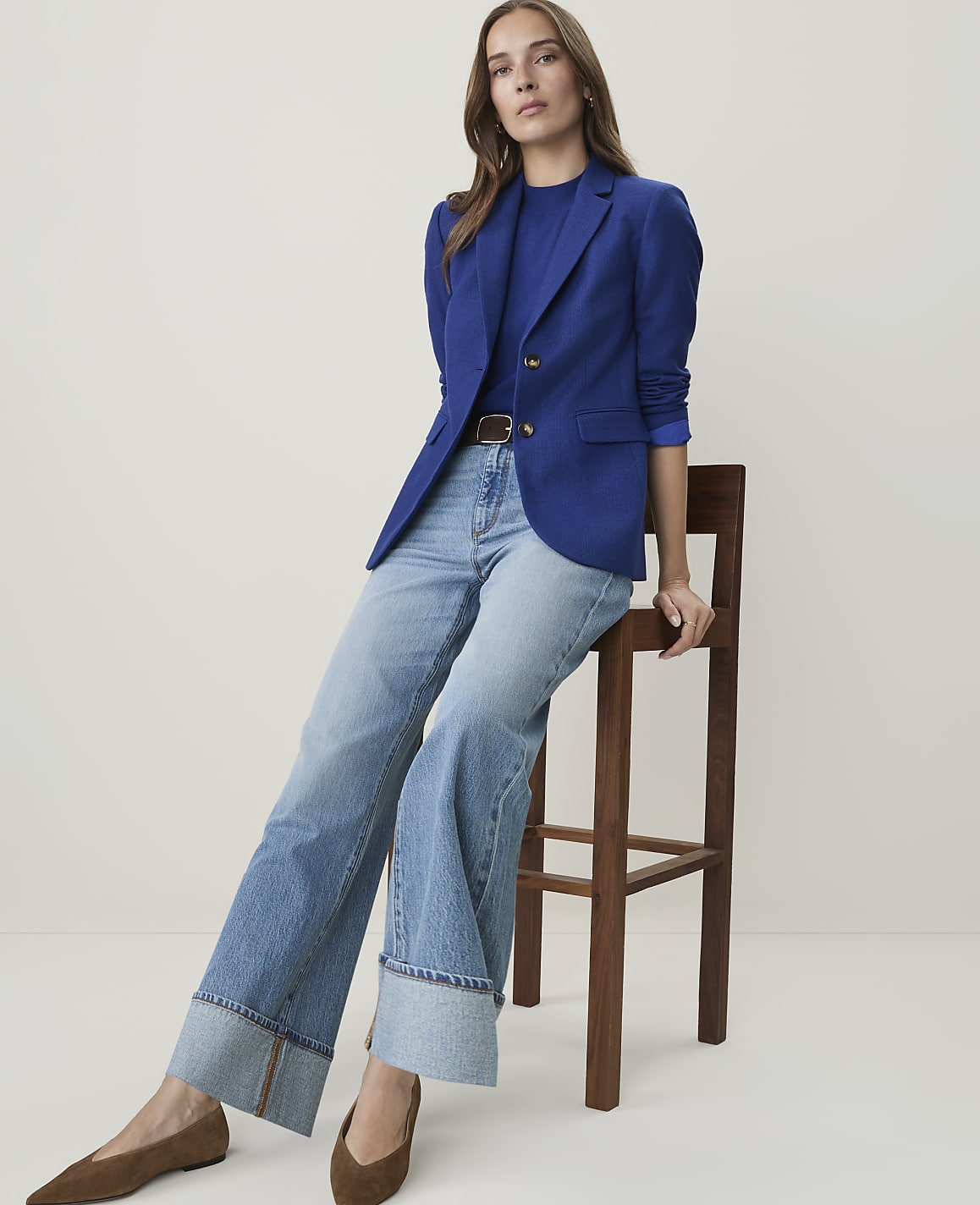 A woman in a blue blazer and wide-leg jeans sits on a wooden stool against a plain white background.