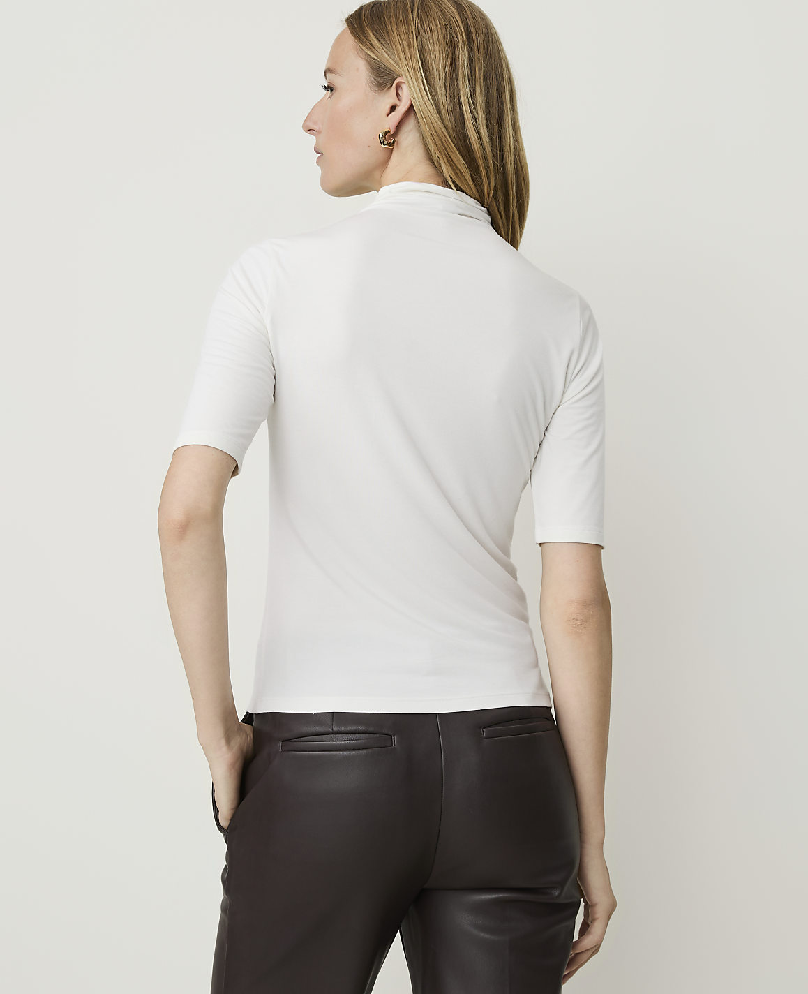 A woman in a white turtleneck shirt and brown leather pants stands against a plain white background.