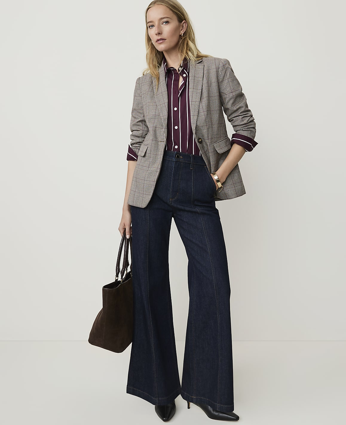 A woman in a patterned blazer and dark wide-leg jeans stands against a plain, light background, holding a brown tote bag.