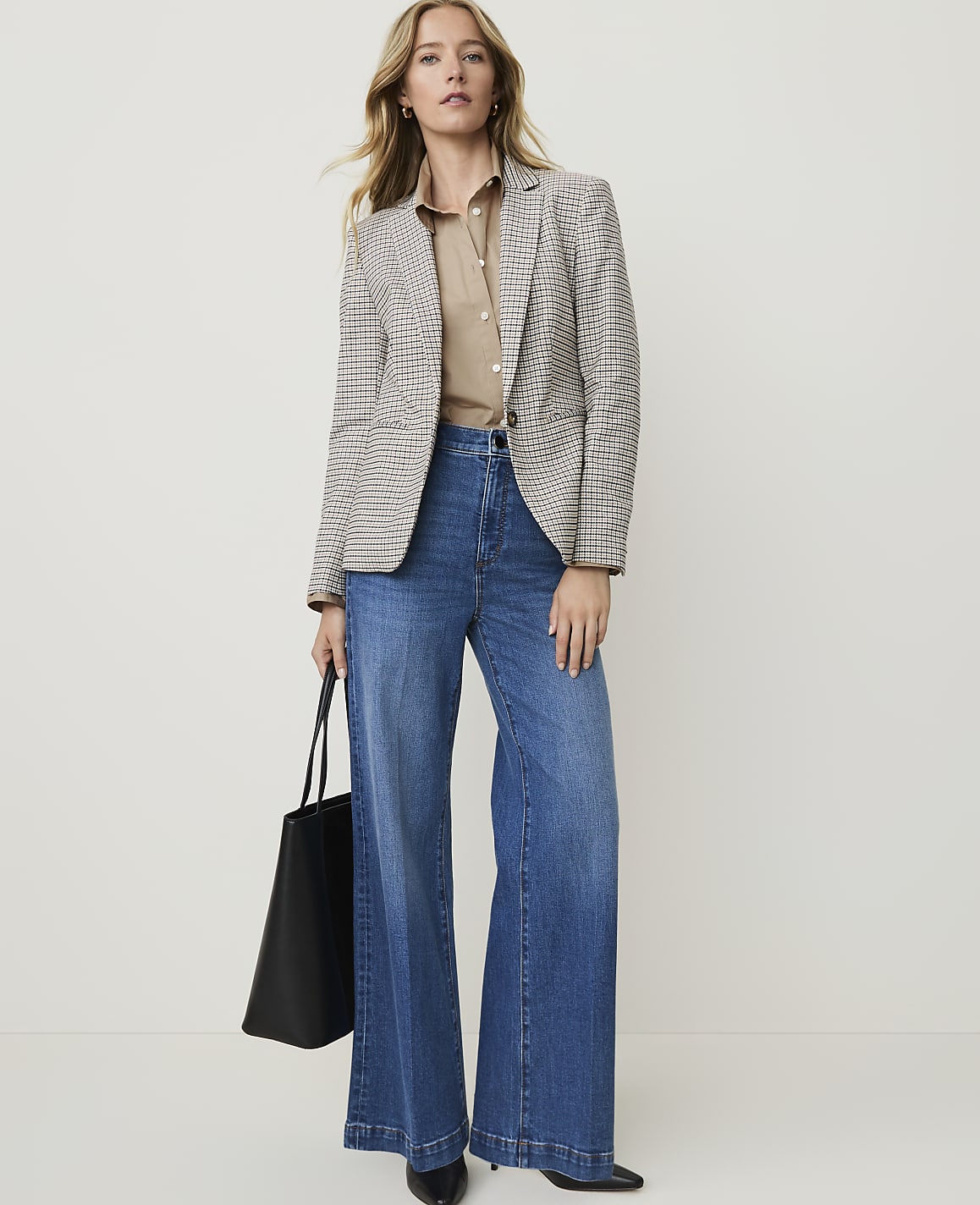A woman in a patterned blazer and wide-leg jeans stands against a plain, light background, holding a black tote bag.