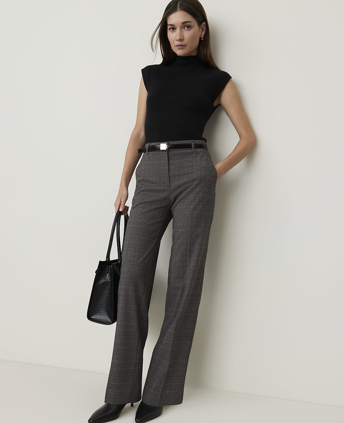 A woman in a black turtleneck and plaid pants stands against a plain white wall, holding a black tote bag.