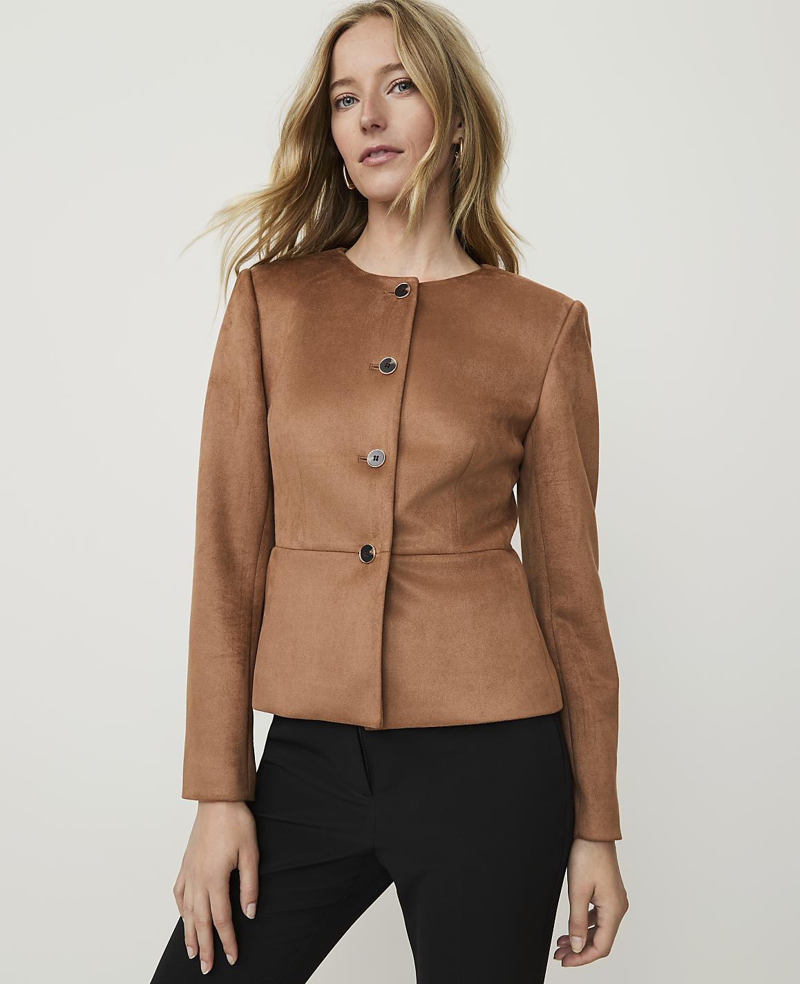 A blonde woman in a brown jacket and black pants stands against a plain, light gray background.