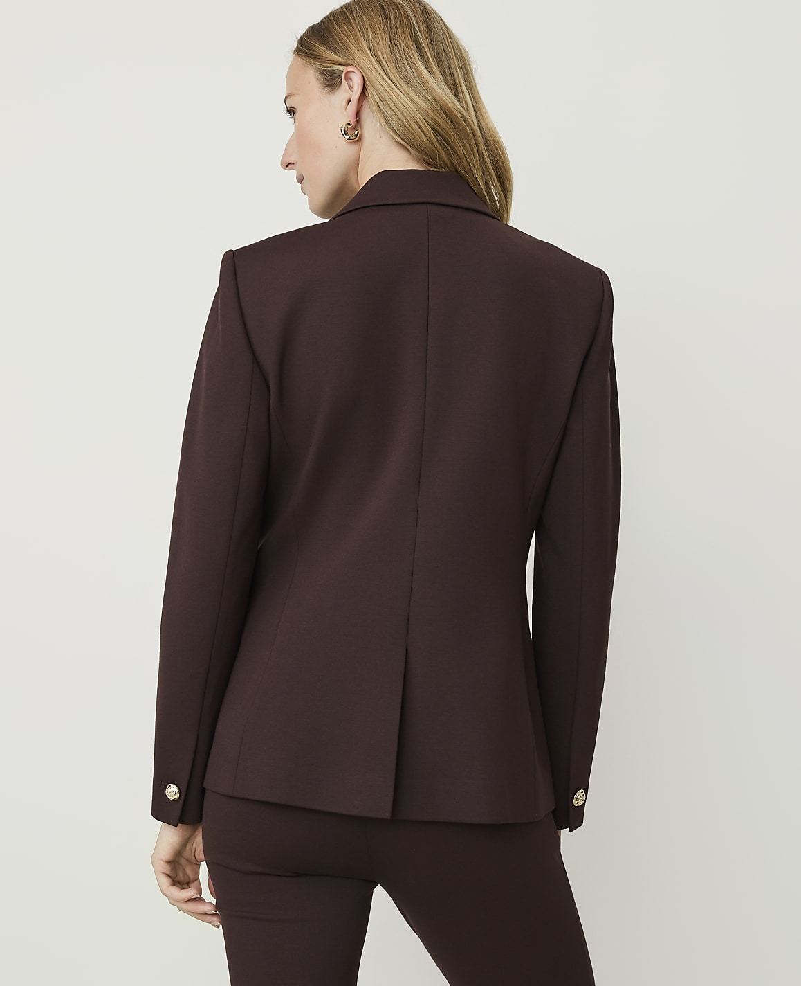 A woman in a brown blazer and pants stands against a plain, light-colored background.